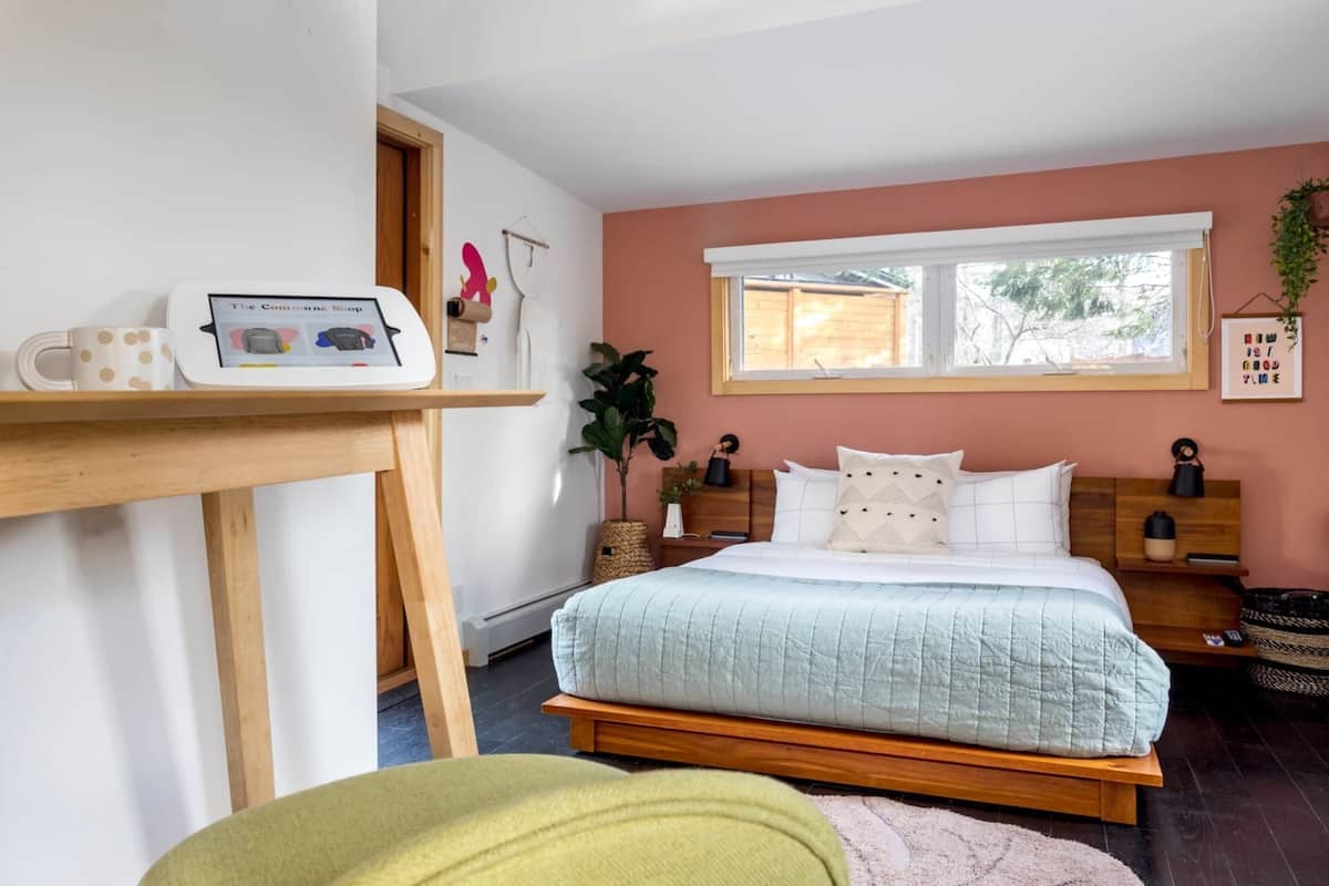 A bedroom with a bed, a bedside table, a window, and a wall-mounted shelf with a mug and a device.