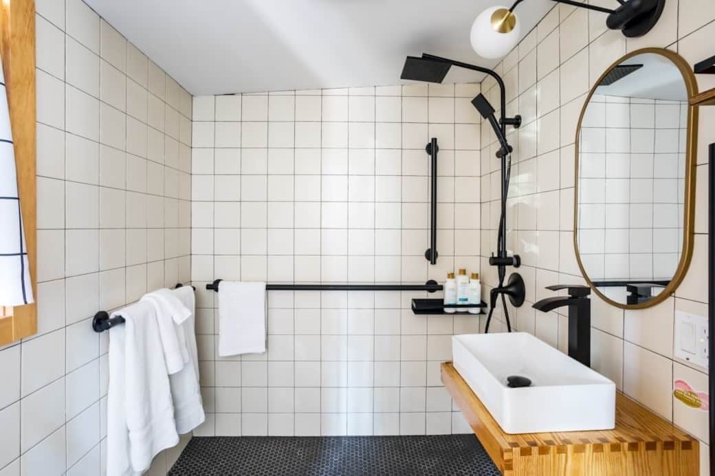 Une salle de bain moderne avec des murs recouverts de carreaux blancs, une pomme de douche noire, un lavabo blanc et un plan de toilette en bois.