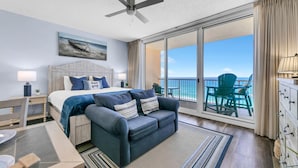 A bedroom with a bed, a sofa, a ceiling fan, a painting, and a balcony with a view of the ocean.