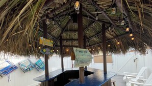 En tiki-bar med ett halmtäckt tak, en bardisk i trä och vita loungestolar.