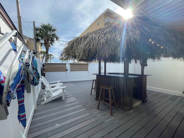 Una terraza de madera con sombrilla de paja, sillas blancas y zona de bar.
