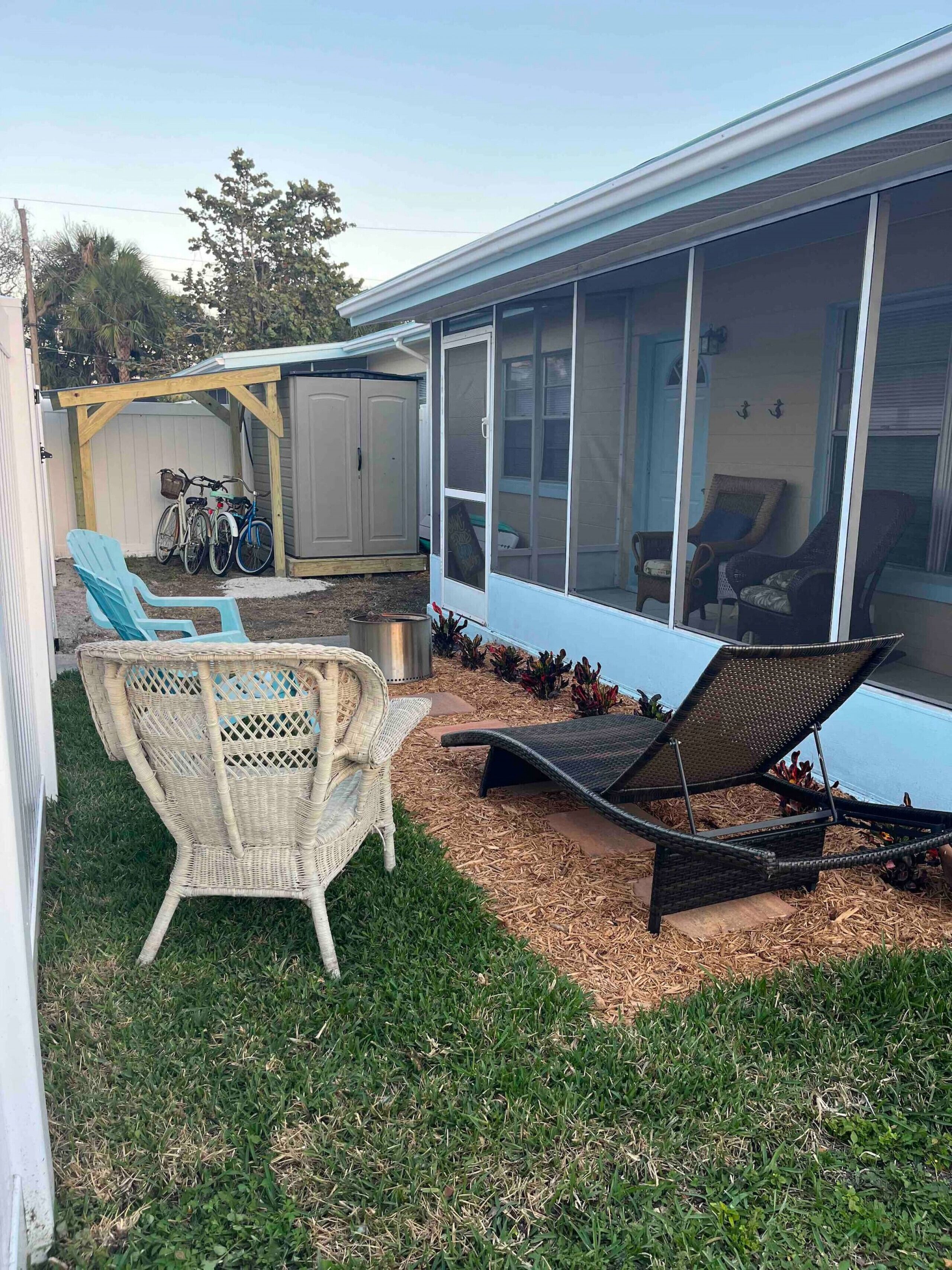 A covered outdoor seating area with wicker chairs and a lounge chair, a covered garage with bicycles, and a house with large windows.