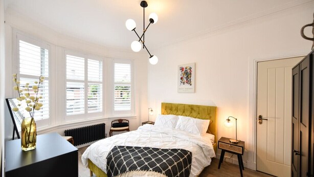 Ein Schlafzimmer mit Bett, Nachttisch, Lampe, Heizkörper und einem Fenster mit Jalousien.