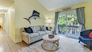 A living room with a sofa, a round coffee table, a lamp, and a view of a balcony labeled "PUBLIC BEACH". - Mermaid Den - Hosted By Moore AI Rentals (Fernandina Beach)