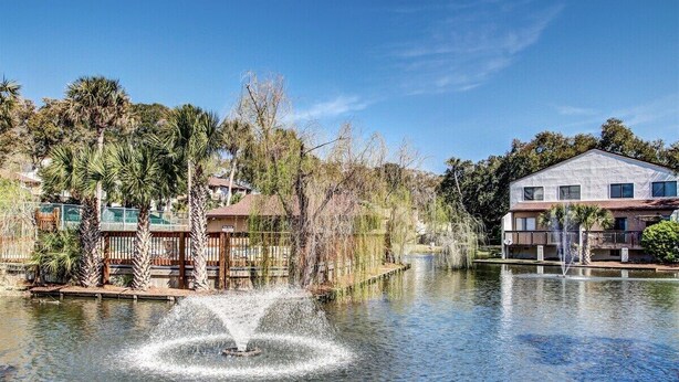 A serene lakeside setting with a fountain, surrounded by palm trees and residential buildings. - Mermaid Den - Hosted By Moore AI Rentals (Fernandina Beach)