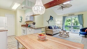 A kitchen with a wooden table, a fruit bowl, a ceiling fan, and a view of the outdoors. - Mermaid Den - Hosted By Moore AI Rentals (Fernandina Beach)