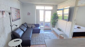 Una sala moderna con un sofá seccional, una mesita redonda y un banco. Hay un televisor montado en la pared, una ventana con vistas al exterior y una puerta que conduce a otra habitación.