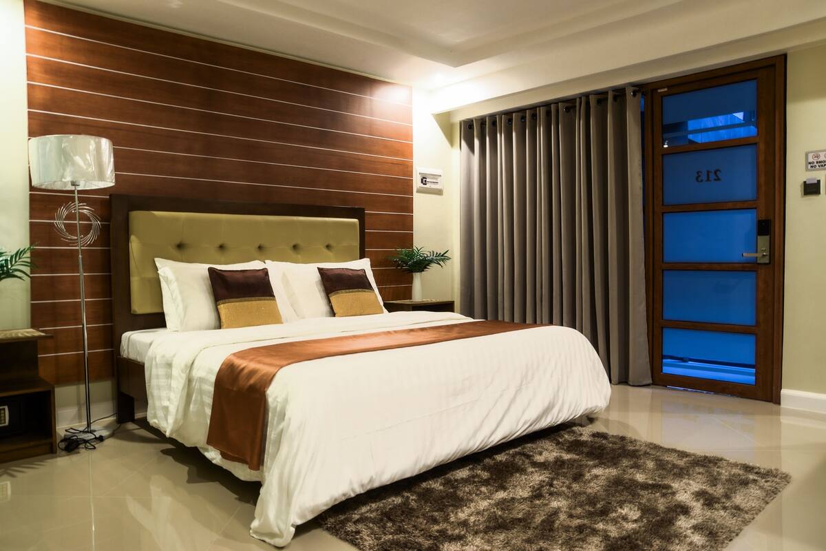 A modern hotel room with a large bed, a wooden headboard, a nightstand, a lamp, and a view of the outside through a glass door.