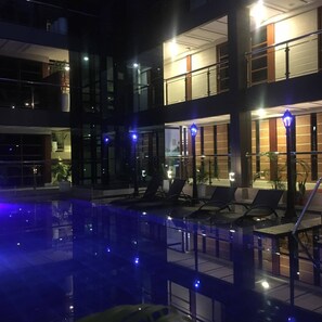 A nighttime view of a hotel with a pool area, lounge chairs, and a modern building facade with large windows. - Panglao Alona Suites (Panglao)