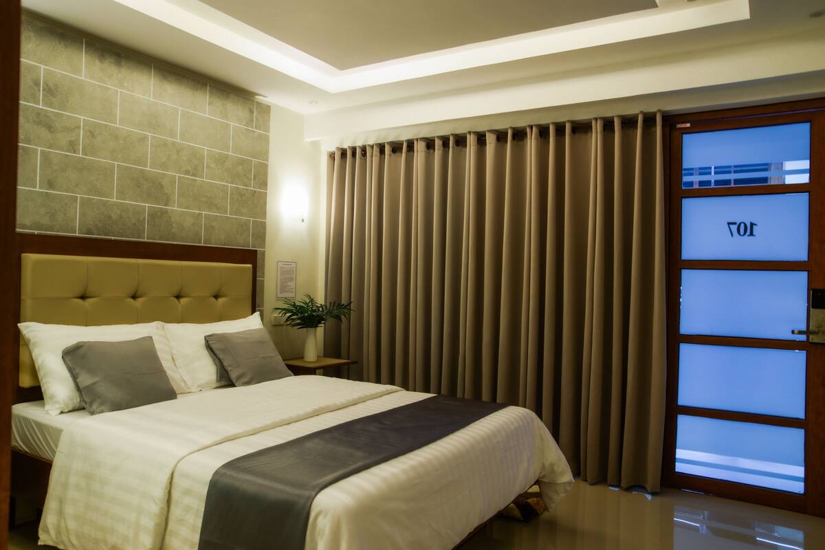 A neatly made bed with a headboard, a bedside table with a plant, and a window with curtains.