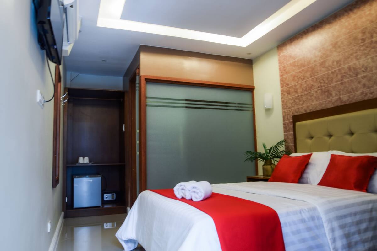 A hotel room with a bed, a red and white bedspread, a white towel on the bed, a nightstand with a plant, and a wardrobe with a mirror.