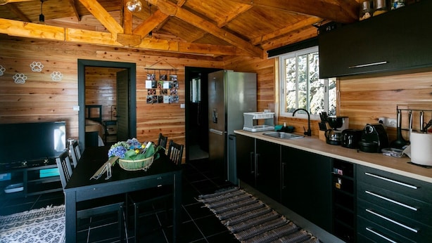 A rustic cabin interior with a wooden ceiling, a dining area with a table and chairs, a kitchen with modern appliances, and a window providing natural light. - Adegada Quinta- Beautiful Cottage in Madalena (Madalena - Pico)