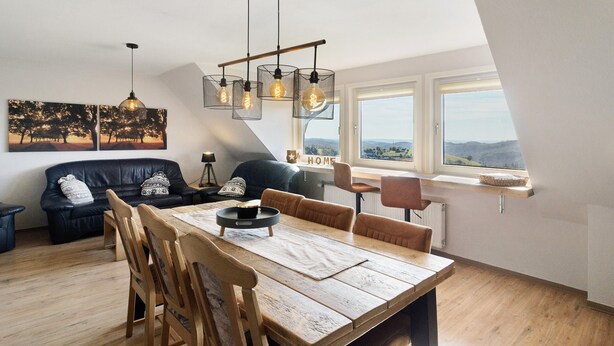 A loft-style room with a dining table, chairs, a sofa, and a view through a large window.