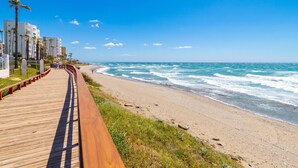 Ein hölzerner Steg führt zu einem Strand mit Meereswellen, Palmen und modernen Gebäuden.