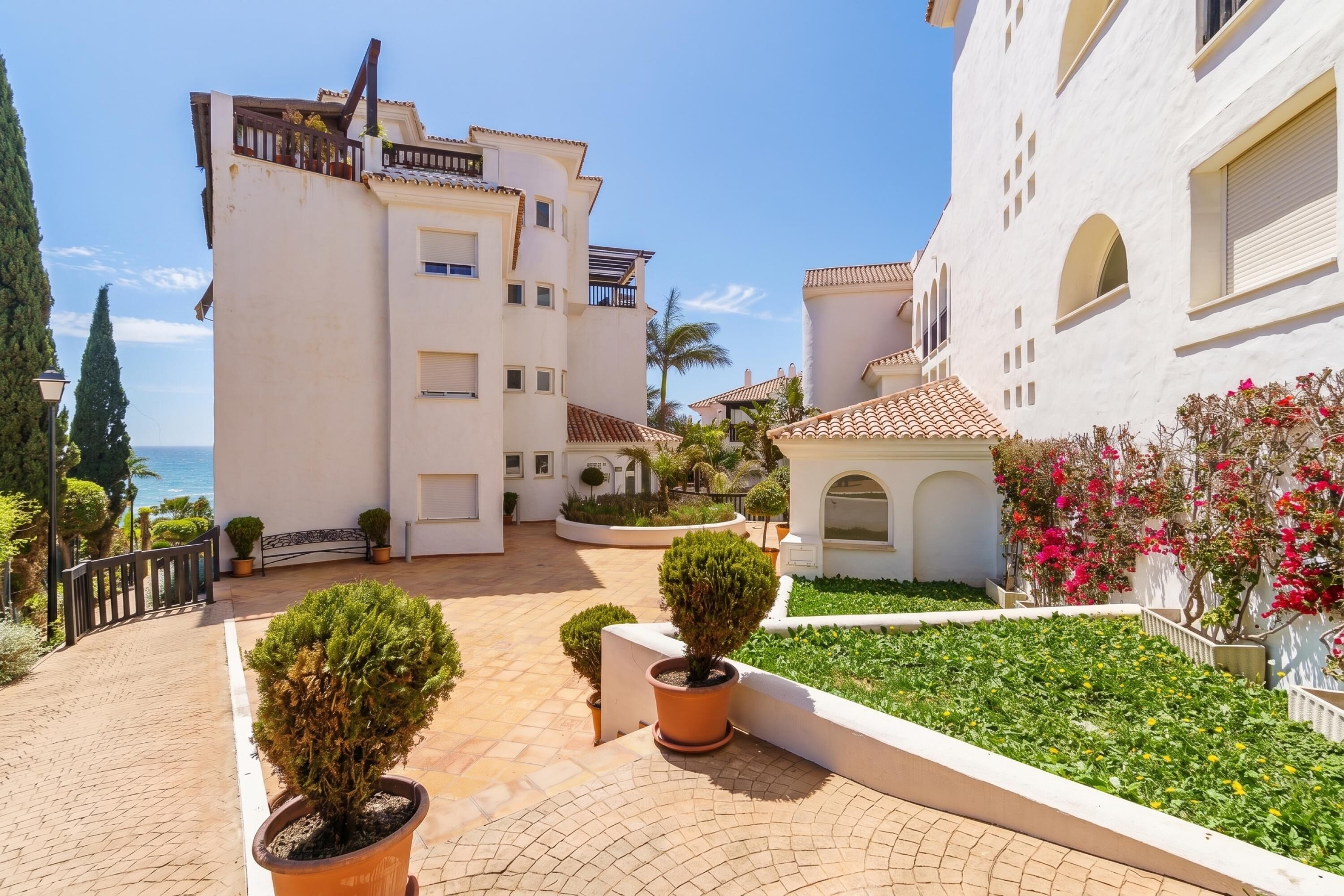 A courtyard with potted plants, a white building with multiple windows, and a view of the sea.
