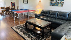 A living room with a ping pong table, a black leather couch, a coffee table, and a floral rug. - Space Coast Retreat: Games, Hot Tub, Fenced yard, Pool Table + Massage Chair (Cocoa)