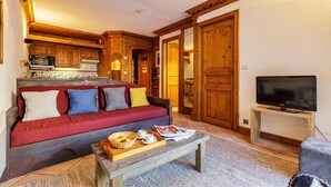 A cozy living area with a red sofa, wooden cabinets, a television, and a kitchen in the background. - Le Cristal GM7 Mountain View, Pool, Private Sauna & Parking, Argentière (Chamonix), France (Argentière (Chamonix))