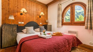 A bedroom with a wooden paneled wall, a large bed with a red quilt, two wall-mounted lamps, a window with a view of greenery, and a wooden nightstand with a lamp. - Le Cristal GM7 Mountain View, Pool, Private Sauna & Parking, Argentière (Chamonix), France (Argentière (Chamonix))