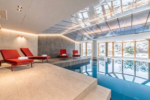 A modern indoor swimming pool with clear glass ceiling, red lounge chairs, and a view of a snowy landscape.