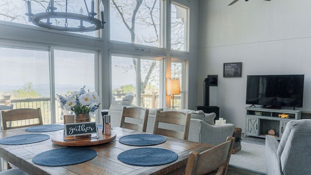 A dining area with a wooden table set for four, a chandelier, and a view through large windows. - The Little Red Barn w/ Spectacular Views & Less than 1 mile - Blue Ridge Parkway (Hillsville)
