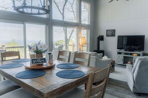 A dining area with a wooden table set for four, a chandelier, and a view through large windows. - The Little Red Barn w/ Spectacular Views & Less than 1 mile - Blue Ridge Parkway (Hillsville)