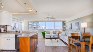 A modern apartment with a kitchen, dining area, and living room. It features a large window, a ceiling fan, and a view of the city.