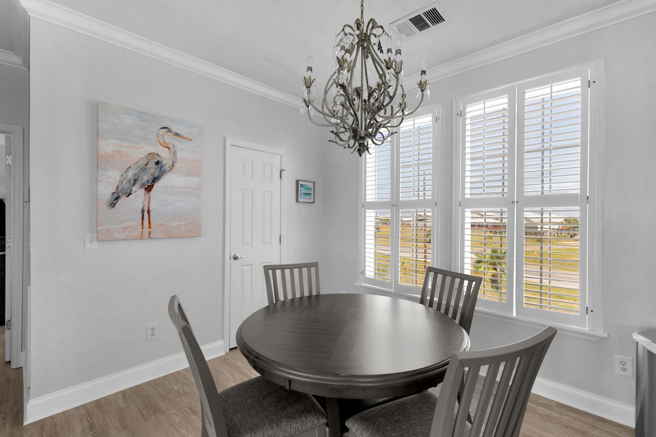 A dining room with a round table, four chairs, a chandelier, a painting of a bird, and a window with blinds.