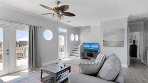 A spacious living room with a grey sofa, a coffee table, and a television. There is a balcony with a view of the ocean and a staircase leading to another level.