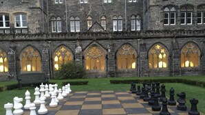 A large outdoor chessboard set up in front of a historic building with arched windows and stone walls.