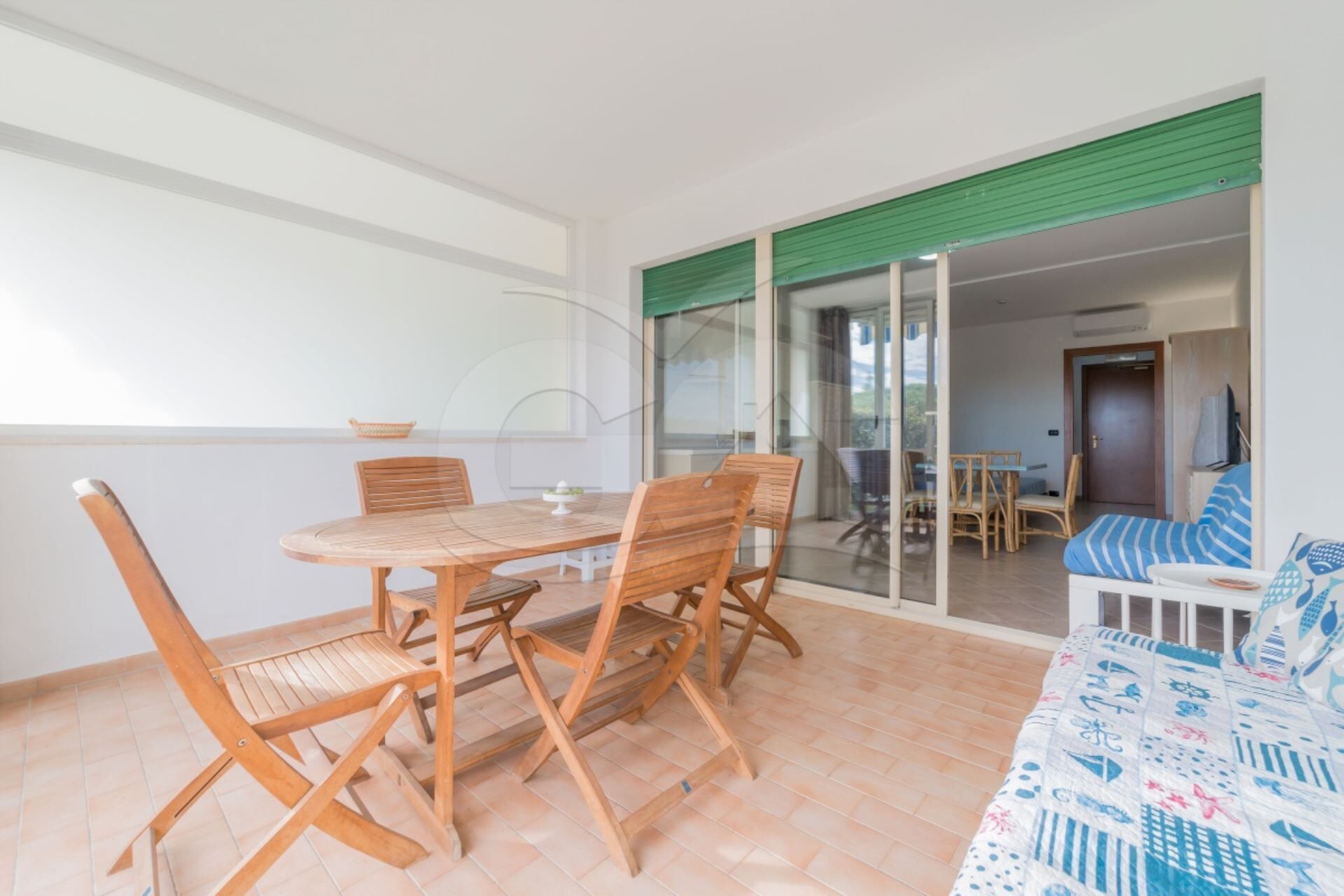 A room with a wooden table and chairs, a sofa with a patterned cover, and a sliding glass door leading to another room with a table and chairs.