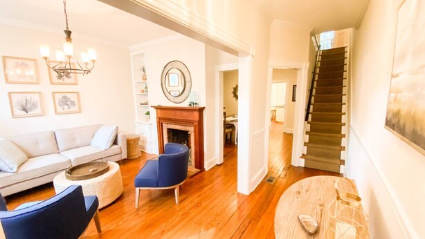 A living room with a sofa, blue armchair, and a round coffee table. There is a fireplace, a staircase, and a wall with framed pictures. - Historic Annapolis Union House with Private Patio & Free Off-Site Parking (Annapolis)