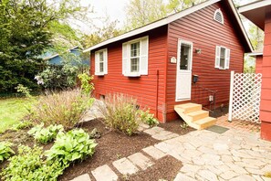 A red wooden house with a front porch, a small garden with various plants, and a white fence.