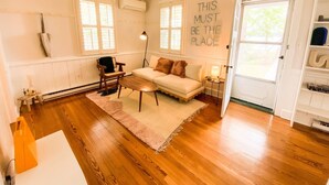 A cozy living room with a wooden floor, a white sofa, a small table, and a chair. There is a large window with blinds, an air conditioning unit, and a door with a shelf.