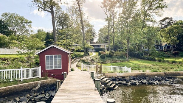A wooden dock leading to a small red cabin by a calm water body, surrounded by lush greenery and a white fence.