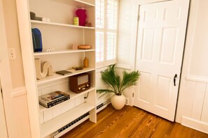 A hallway with white walls, a white door, and a wooden floor. Shelves are stocked with books, a radio, and decorative items.