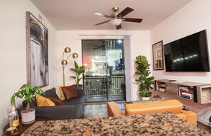 A living room with a grey sofa, orange ottoman, and a TV on the wall. There is a ceiling fan, a plant, and a balcony with a glass door. - Luxury 1BR in the heart of The Domain - walk to dining, shopping, & nightlife. (Austin)