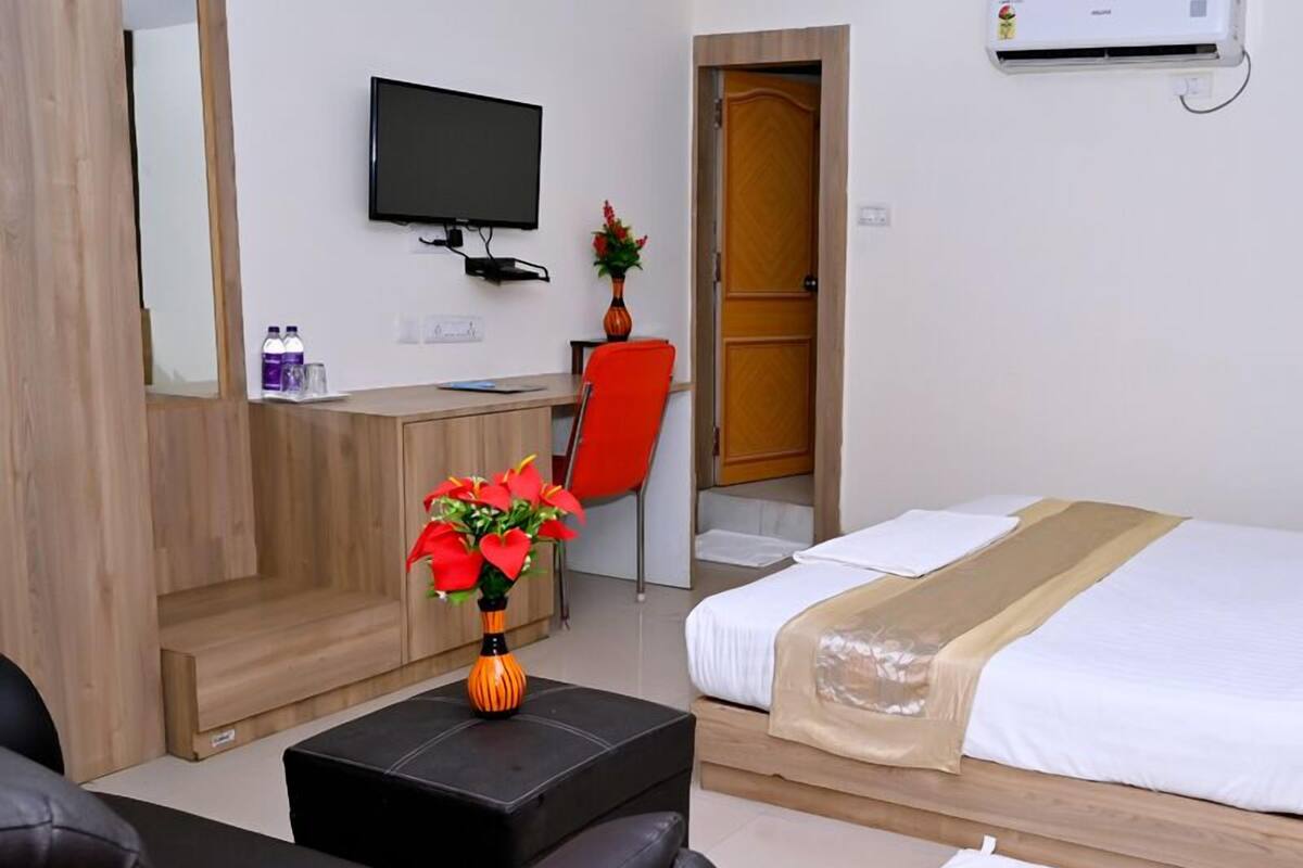 A hotel room with a bed, a desk, a chair, a TV, and a small table with a vase of flowers.