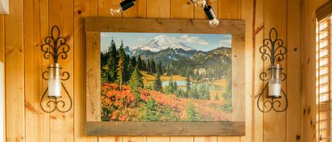 A wooden interior with a dining table, benches, and a wall-mounted painting of a mountain landscape.