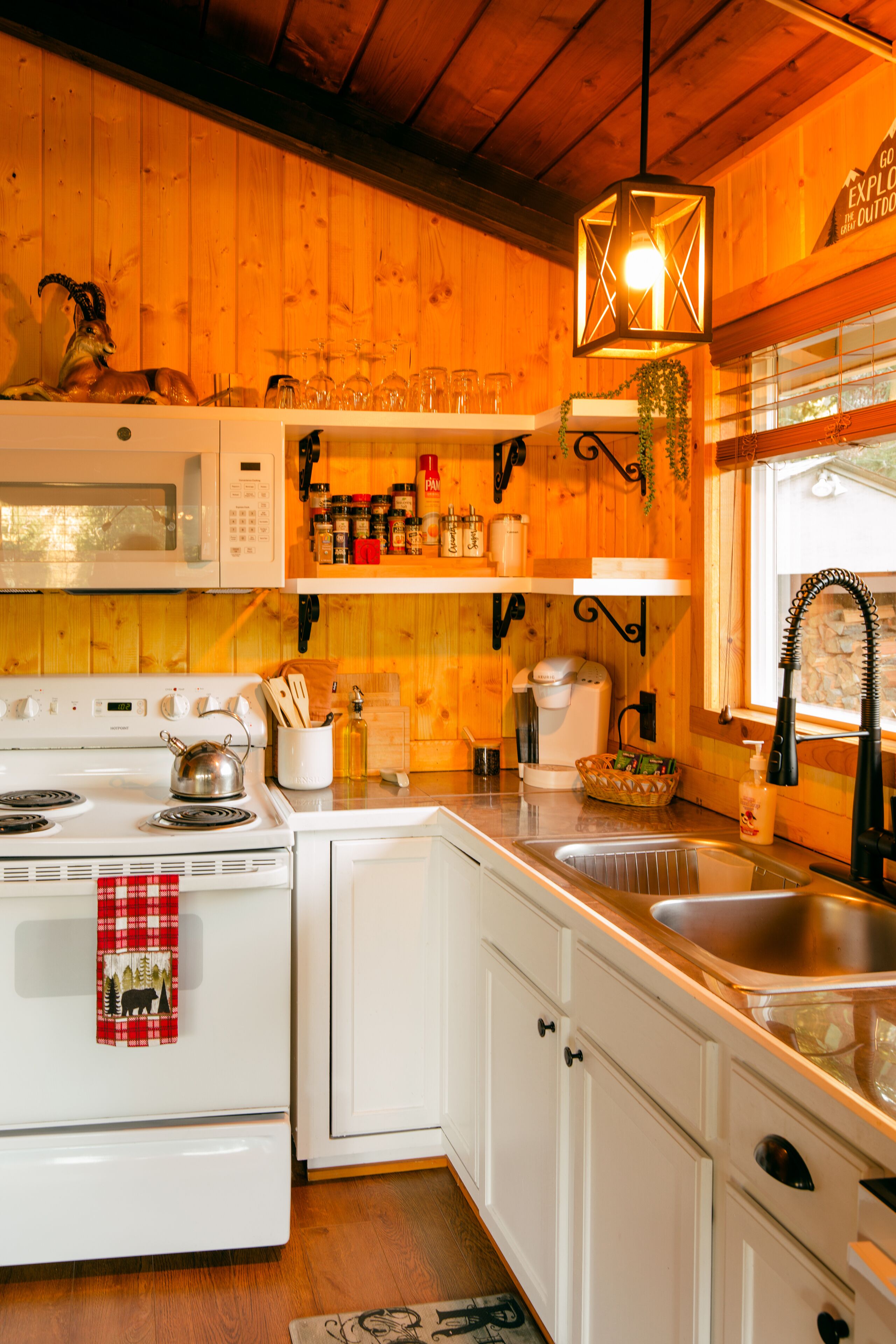 Una cocina con armarios blancos, una estufa, un microondas y un lavabo. Hay repisas con frascos y una lámpara colgante.