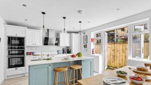 A modern kitchen with white cabinets, a blue island, wooden stools, and a dining table set with plates and cutlery.