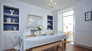 Un coin repas avec un banc en bois, des chaises blanches, une table recouverte d’une nappe à motifs, un lustre et une bibliothèque agrémentée d’objets décoratifs.