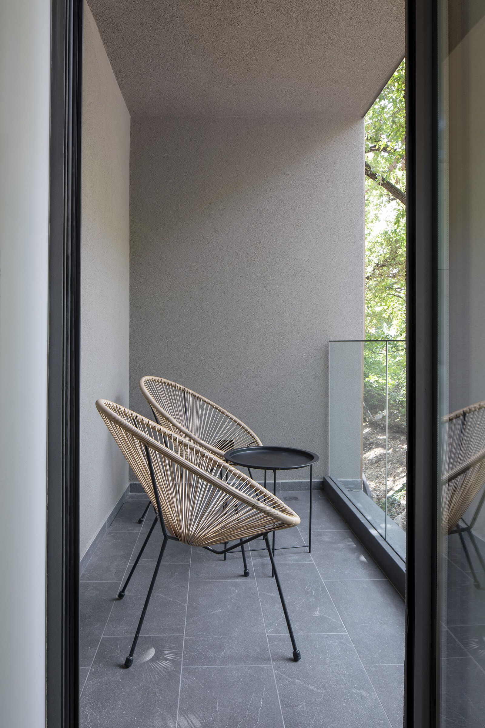 A balcony with two wicker chairs and a small table, a glass railing, and a view of trees.