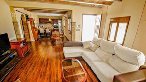 A living room with a sofa, a glass coffee table, and a TV. There is a kitchen in the background.