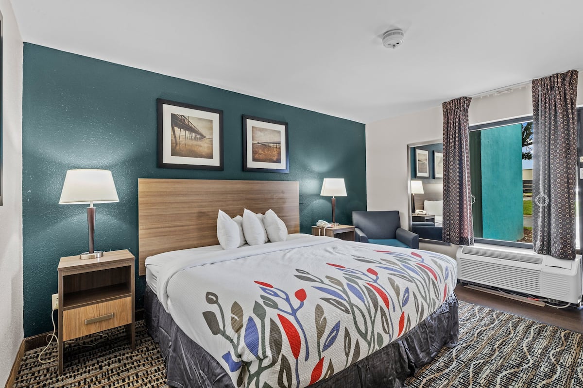 A hotel room with a bed, two bedside lamps, a nightstand, a chair, a window with curtains, and two framed pictures on the wall.