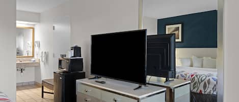Ett hotellrum med en säng, ett skrivbord med en platt-TV, ett litet kylskåp, en mikrovågsugn, en spegel och ett badrum som syns i bakgrunden.