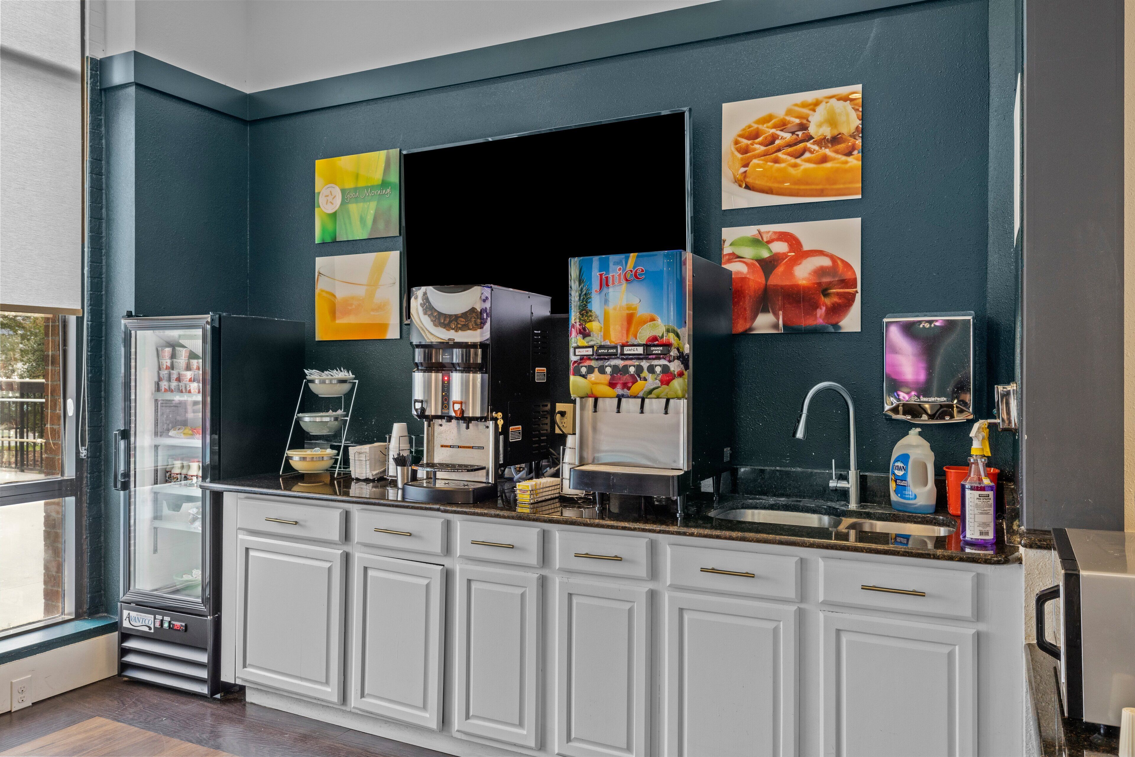 A kitchen with a refrigerator, coffee machine, juice dispenser, and a sink.