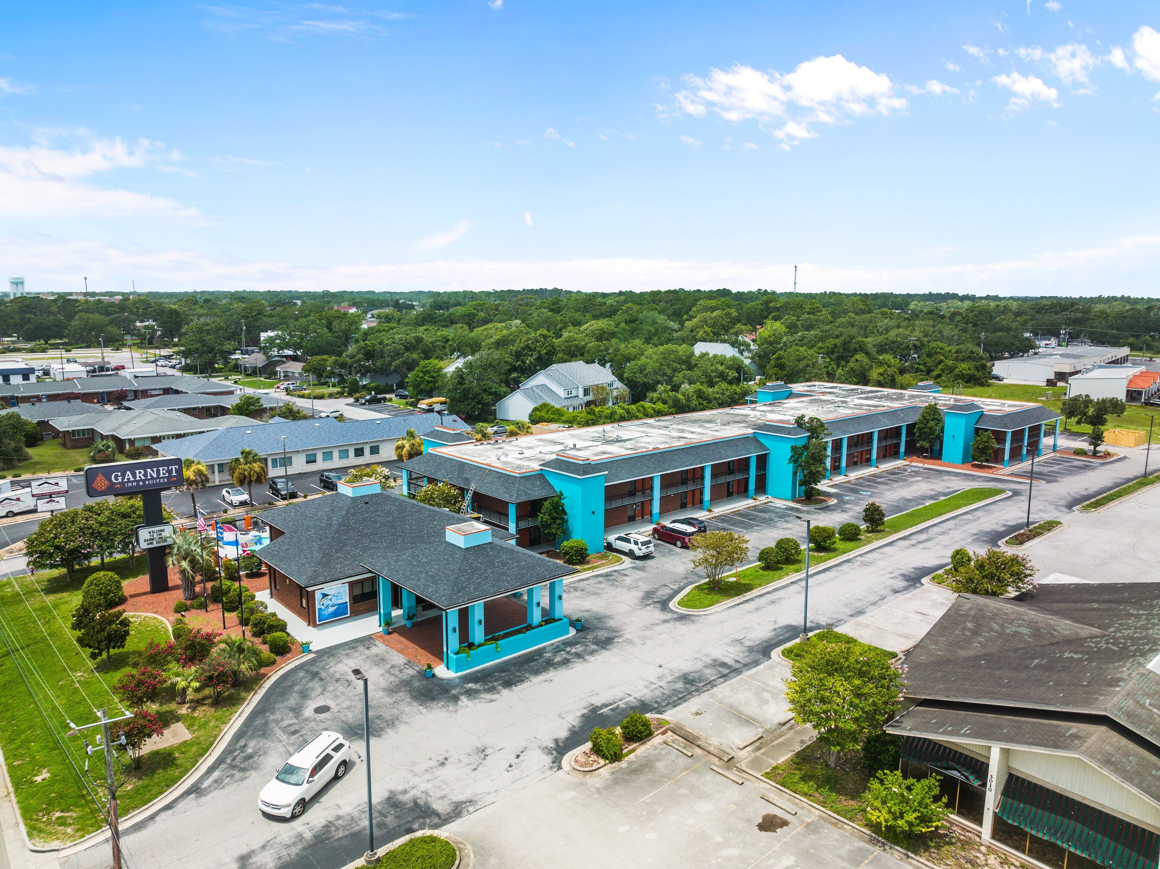 Um hotel de vários andares com esquema de cores azul e branco, vários estacionamentos e paisagem verde ao redor.