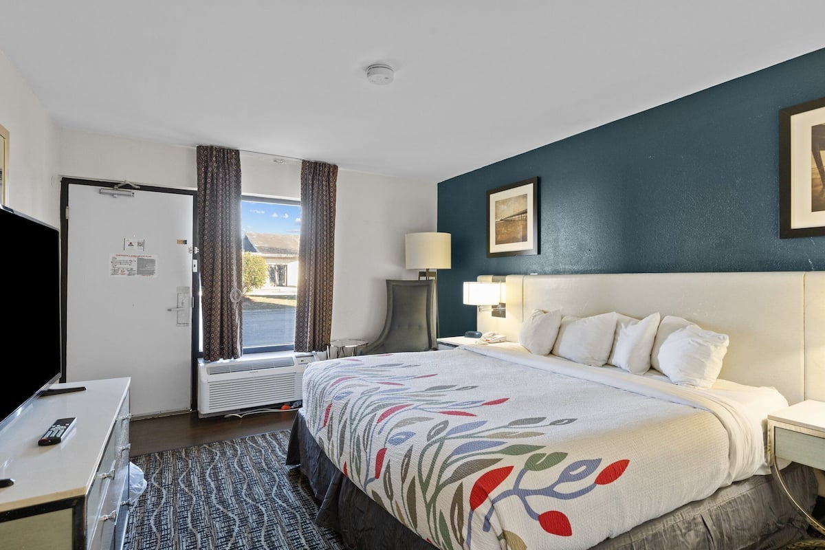 A hotel room with a bed, a desk, a chair, a lamp, and a window with curtains.