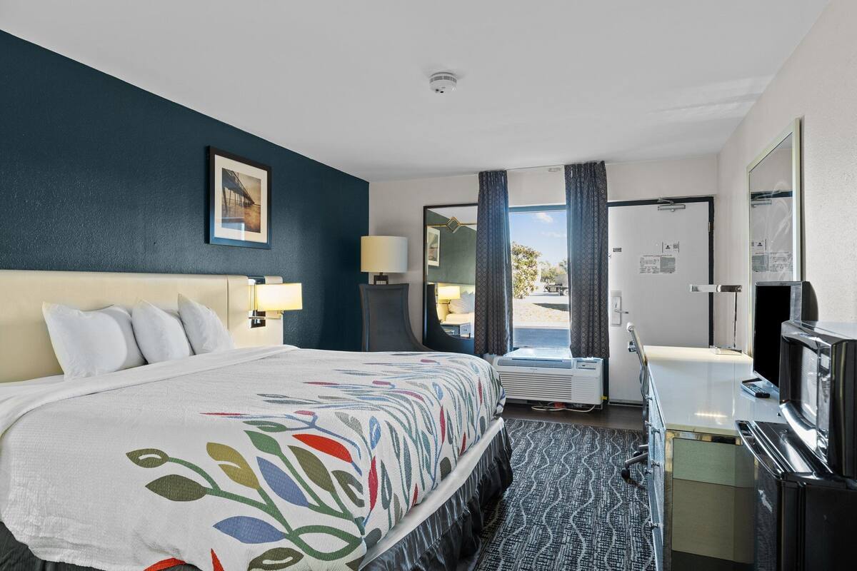 A hotel room with a large bed, a desk with a computer, a mirror, and a window with curtains.