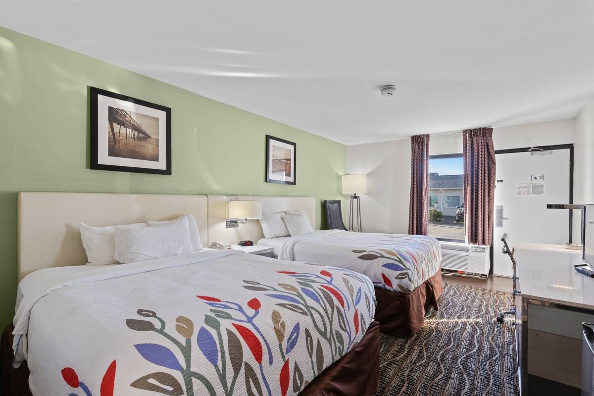 A hotel room with two beds, a desk, a chair, a window with curtains, and a picture on the wall.